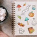Photograph of a sketchbook filled with autumn-themed drawings. Next to the sketchbook is a blanket and a cup full of marshmallows.